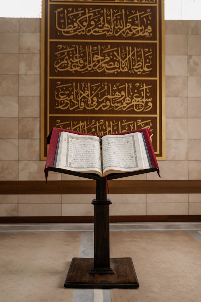 A Quran open on a stand within a mosque interior, featuring Arabic calligraphy on the wall.